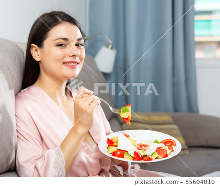 Satisfied brunette sitting on a sofa and eating salad Satisfied brunette sitting on a sofa and eating salad 85604010