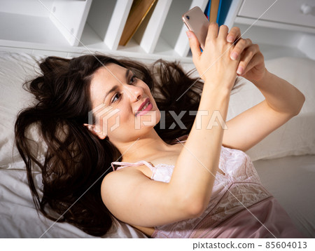 Young sexy woman lying and using phone on white sheet Young sexy woman lying and using phone on white sheet 85604013