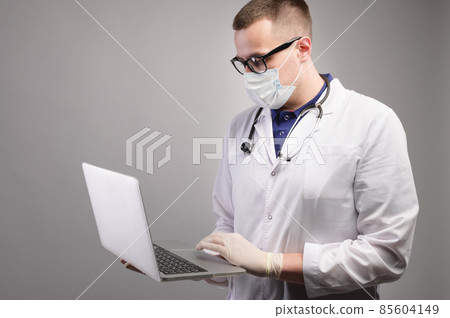 Successful young caucasian male doctor in a white medical uniform and with a stethoscope, busy working on a laptop, typing on a computer, consulting a patient online. Studio portrait Successful young caucasian male doctor in a white medical uniform and with a stethoscope, busy working on a laptop, typing on a computer, consulting a patient online. Studio portrait 85604149