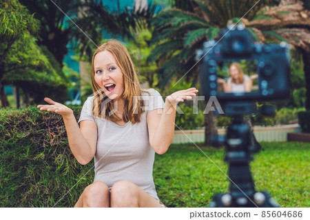 A young female blogger is leading her video blog in front of the camera in the park. Blogger concept A young female blogger is leading her video blog in front of the camera in the park. Blogger concept 85604686