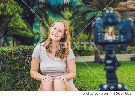 A young female blogger is leading her video blog in front of the camera in the park. Blogger concept A young female blogger is leading her video blog in front of the camera in the park. Blogger concept 85604692