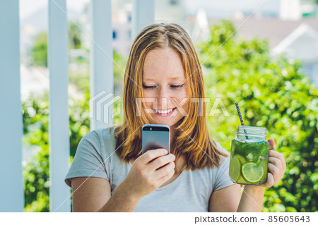 A young woman holds a mason jar in her hand with a mojito and a smartphone. Spring drink concept A young woman holds a mason jar in her hand with a mojito and a smartphone. Spring drink concept 85605643