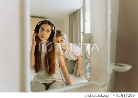 Teenage girl in headphones in front of a mirror, little sister interferes, grimaces. 85617299