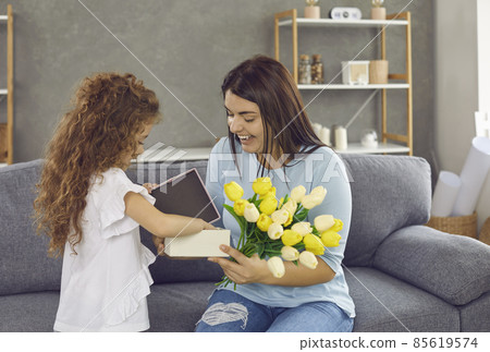 Lifestyle moment. Little daughter congratulates her loving mother on holiday by giving gifts and flowers. Happy young excited woman who is delighted with gift looks at him sitting at home on couch. Lifestyle moment. Little daughter congratulates her loving mother on holiday by giving gifts and flowers. Happy young excited woman who is delighted with gift looks at him sitting at home on couch. 85619574