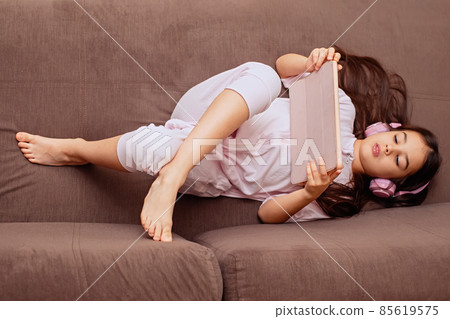 A little cute girl in pink home clothes and pink headphones, lies on the couch with digital tablet 85619575