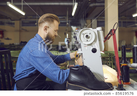 Profile portrait of shoe factory worker in workshop. Side view serious man sitting at table and using industrial sewing machine to make new black leather boots. Footwear manufacturing industry concept 85619576