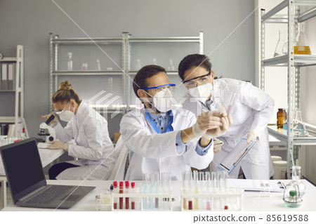 Scientists doing research in science laboratory. Two medical chemists in white lab coats and glasses standing at table with laptop computer looking at sample liquids and discussing future experiment Scientists doing research in science laboratory. Two medical chemists in white lab coats and glasses standing at table with laptop computer looking at sample liquids and discussing future experiment 85619588