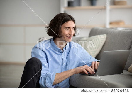 Blogging concept. Young guy using laptop, writing post to social network while sitting on floor near couch, free space Blogging concept. Young guy using laptop, writing post to social network while sitting on floor near couch, free space 85620498