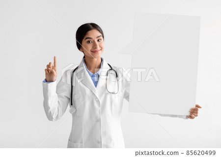 Smiling young indian female doctor in uniform show finger up and hold big banner Smiling young indian female doctor in uniform show finger up and hold big banner 85620499