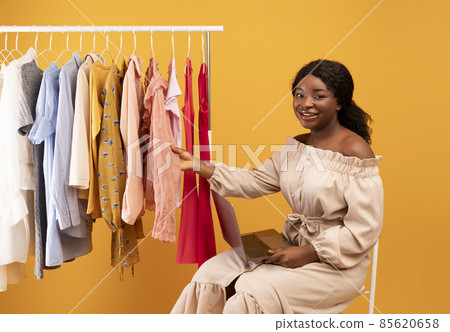 Young black woman with laptop touching clothes on rack, shopping online for new outfits, purchasing on internet 85620658