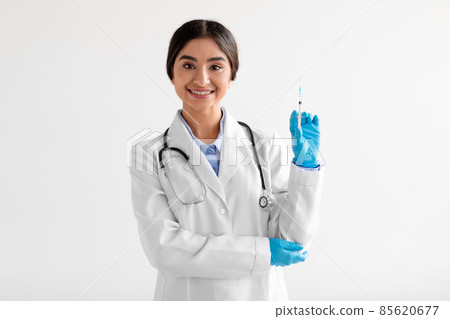 Smiling young hindu female medical worker in uniform, protective gloves, shows syringe with vaccine 85620677