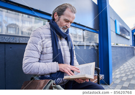 Mature man reading book while waiting for public transportation Mature man reading book while waiting for public transportation 85620768