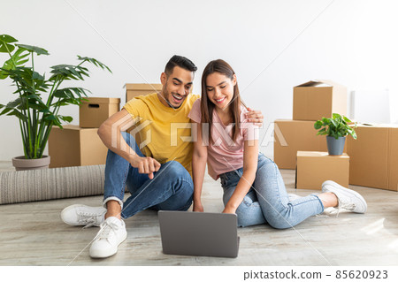 Moving day. Young diverse couple with laptop purchasing household goods for new home online, sitting on floor with pc Moving day. Young diverse couple with laptop purchasing household goods for new home online, sitting on floor with pc 85620923