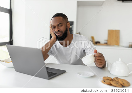 Tired black man using laptop sitting at kitchen table Tired black man using laptop sitting at kitchen table 85621014