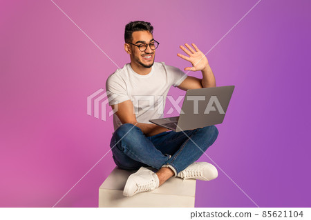 Young Arab guy sitting cross legged with laptop, waving at webcam, communicating to friend or colleague in neon light Young Arab guy sitting cross legged with laptop, waving at webcam, communicating to friend or colleague in neon light 85621104