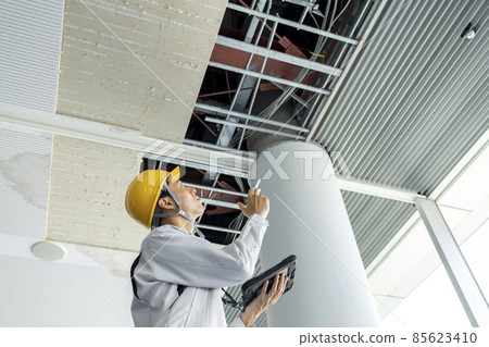A man in work clothes holding a tablet and investigating the damaged part of the building ceiling 85623410