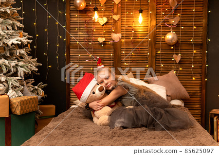 A girl hugs a bear toy in a Christmas cap bear Christmas kid child little, from people home for play sitting room, small joyful. Bedroom bed holding, background wake 85625097