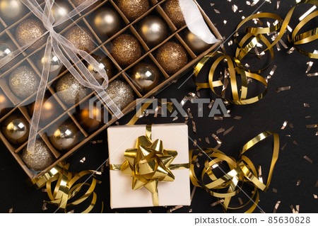 Holiday gift box or present with ribbon, golden confetti and gold baubles on black background. Magic New Year greeting card. Christmas Decoration. Mock up. Flat lay.  85630828
