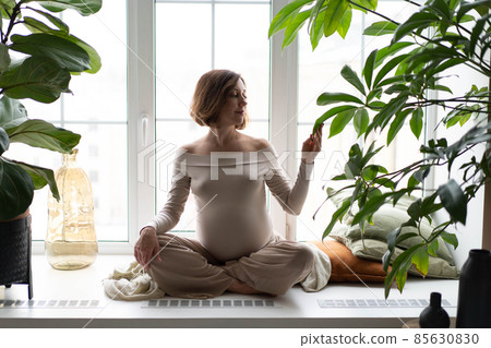 Beautiful young pregnant woman is sitting on the window. Meditation and calm relaxation. Concept of online classes, quarantine, online classes at home during a pandemic. 85630830