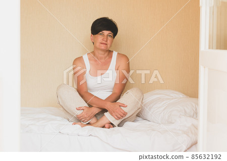 A middle-aged woman in casual home clothes has woken up and is sitting cross-legged on the bed. Human psychology 85632192
