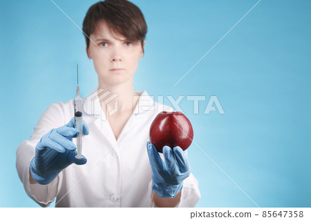Portrait of a female doctor standing with an Apple and a syringe on a blue background. concept of enhancing the growth of vegetables and fruits 85647358