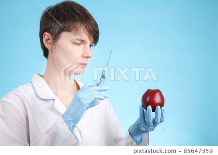 Portrait of a female doctor standing with an Apple and a syringe on blue as idea of antibiotics in vegetables and fruits 85647359