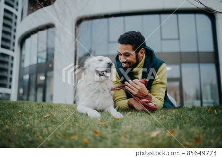 Happy young man lying on grass with his dog outdoors in town. Happy young man lying on grass with his dog outdoors in town. 85649773