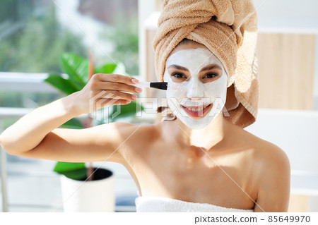 Beautiful young woman with towel wrapped around her head applying face mask at the bathroom 85649970