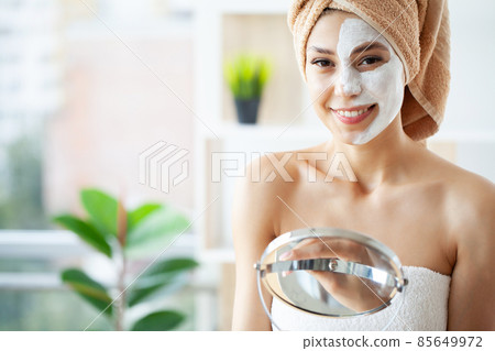 Beautiful young woman with towel wrapped around her head applying face mask at the bathroom 85649972