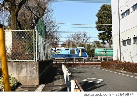 A freight train that can be seen at the end of the road. The blue of the electric locomotive shines in the white building and the blue sky. 85649974