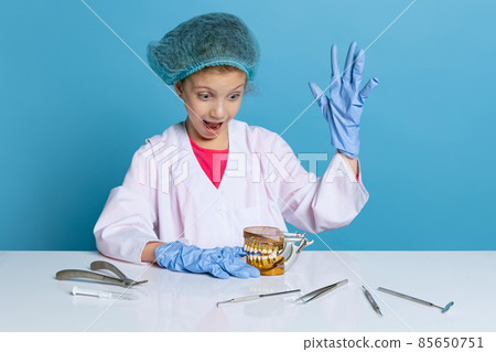 Emotional cute little girl, child in image of dentist doctor wearing white lab coat and gloves isolated on blue studio background Emotional cute little girl, child in image of dentist doctor wearing white lab coat and gloves isolated on blue studio background 85650751
