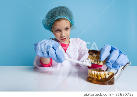 Emotional cute little girl, child in image of dentist doctor wearing white lab coat and gloves isolated on blue studio background 85650752