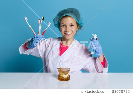Emotional cute little girl, child in image of dentist doctor wearing white lab coat and gloves isolated on blue studio background 85650753
