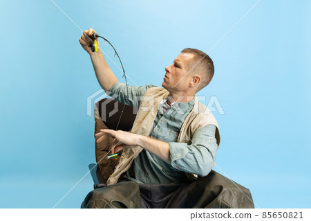 Serious young man, fisherman with fishing accessories sitting on chair isolated over blue studio background Serious young man, fisherman with fishing accessories sitting on chair isolated over blue studio background 85650821