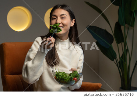 Cheerful young Asian woman who eats fresh salad and smiles sitting on a chair in the living room Cheerful young Asian woman who eats fresh salad and smiles sitting on a chair in the living room 85656716