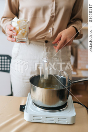 Vertical front view of female artisan adding white dry soy wax in glass jar into pot of boiling water for creating candle building mixture. Vertical front view of female artisan adding white dry soy wax in glass jar into pot of boiling water for creating candle building mixture. 85658117