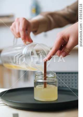 Close-up vertical shot of unrecognizable female artisan pouring melted wax into tranparent glass jar with wick at home. Close-up vertical shot of unrecognizable female artisan pouring melted wax into tranparent glass jar with wick at home. 85658118