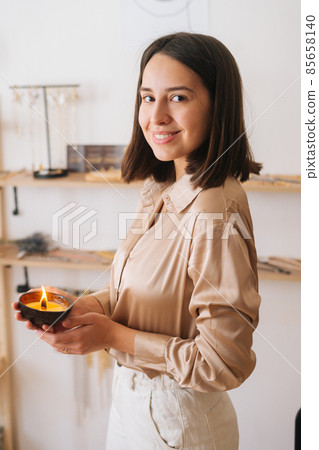 Vertical portrait of smiling young woman holding burning scented handmade candle in palms, looking at camera. Vertical portrait of smiling young woman holding burning scented handmade candle in palms, looking at camera. 85658140