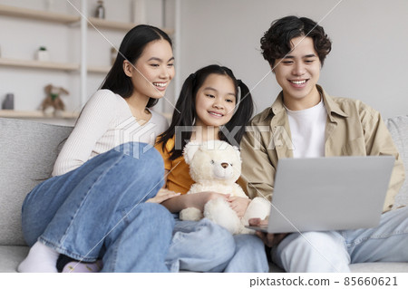 Cheerful young chinese male, female and teen daughter with toy watch video on laptop, searching, sits on sofa Cheerful young chinese male, female and teen daughter with toy watch video on laptop, searching, sits on sofa 85660621