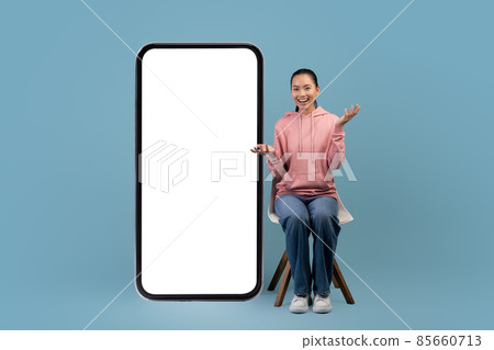 Surprised asian woman sitting on chair by big smartphone, mockup 85660713