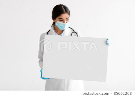 Young indian woman doctor in uniform, gloves and protective mask shows banner with blank space Young indian woman doctor in uniform, gloves and protective mask shows banner with blank space 85661268