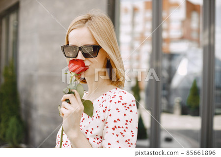 Young blonde woman standing near cafe and holding one rose Young blonde woman standing near cafe and holding one rose 85661896
