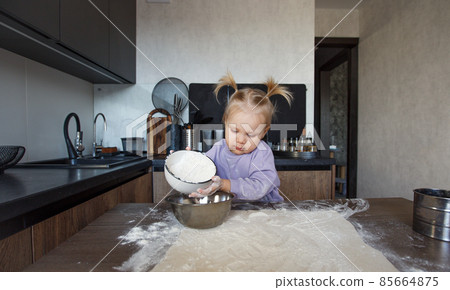 a beautiful little girl is learning to cook in the kitchen. The child spills flour on the kitchen table 85664875