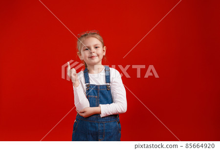a little beautiful schoolgirl in a denim dress poses on a red isolated background. a little beautiful schoolgirl in a denim dress poses on a red isolated background. 85664920