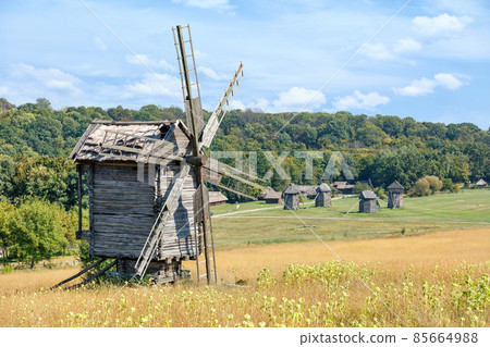 Antique wooden windmill against the backdrop of a rural antique summer landscape with blue sky and dense forest. 85664988