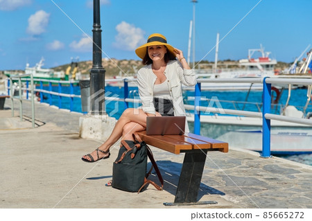 Mature woman in a hat with a laptop on the sea promenade in the city. Mature woman in a hat with a laptop on the sea promenade in the city. 85665272