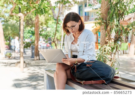 Middle-aged serious woman typing text on laptop outdoors 85665273