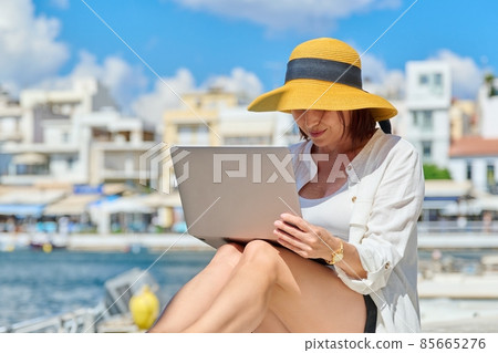 Mature woman in hat with laptop on sea promenade in city Mature woman in hat with laptop on sea promenade in city 85665276