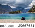 Popular Oludeniz beach on the background of the mountains in pre-thunderstorm weather. There are a couple of people sitting on the pebble shore. Oludeniz, Mugla, Turkey. 85667681