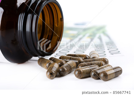 Close-up of a prescription bottle and drug capsules lying on a white background with one hundred euro bills in the background. 85667741